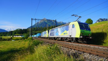 Zug von Cargobeamer mit Sattelaufliegern und Containern fährt vor Alpenpanorama