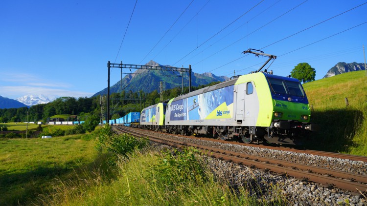 Zug von Cargobeamer mit Sattelaufliegern und Containern fährt vor Alpenpanorama