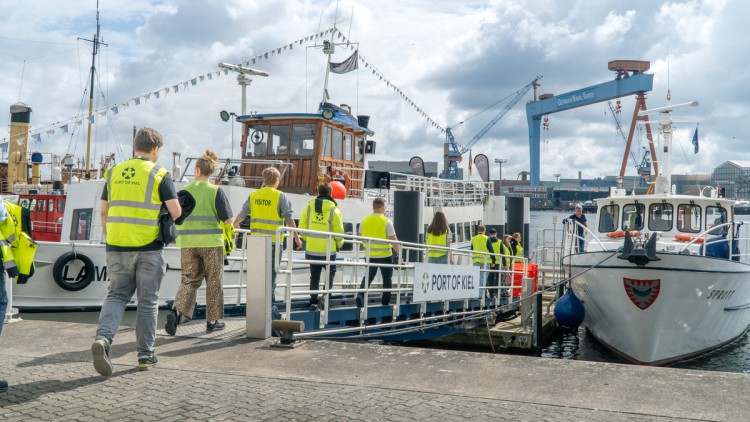 Azubis laufen am Hafen Kiel in gelben Warnwesten nacheinander über einen Steg auf ein Boot