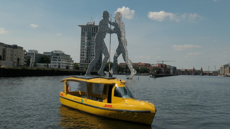 Eine kleine Fähre in gelber DHL-Lackierung vor den "Molecule Men" im Osthafen